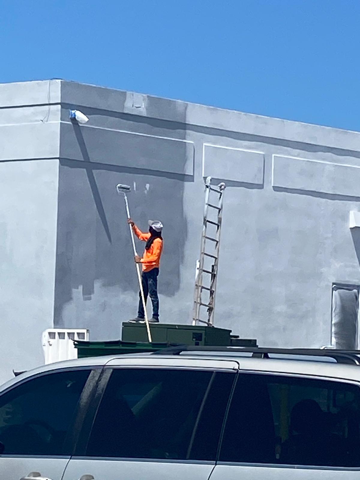 Painter in orange safety vest using an extension roller to apply gray paint to a commercial parapet wall in Doral