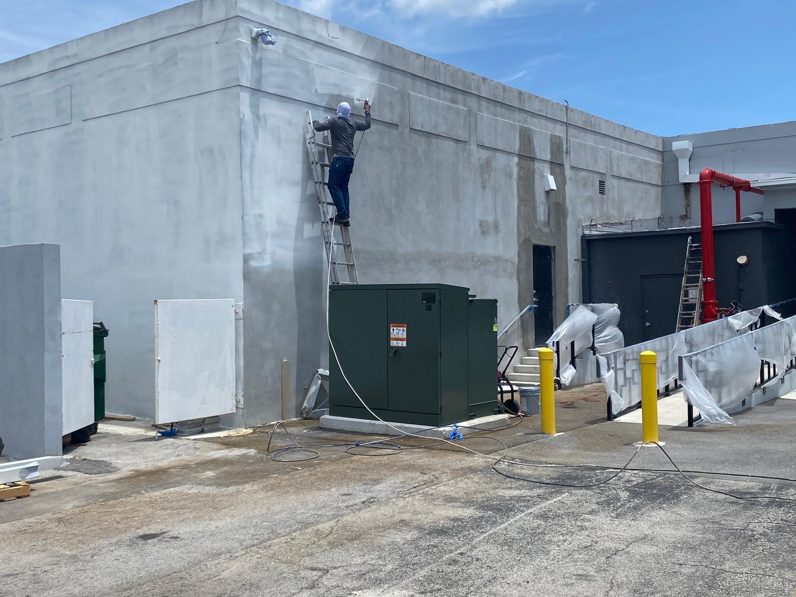 Painter on ladder spray-painting the gray stucco exterior wall of an industrial commercial building in Hialeah Florida