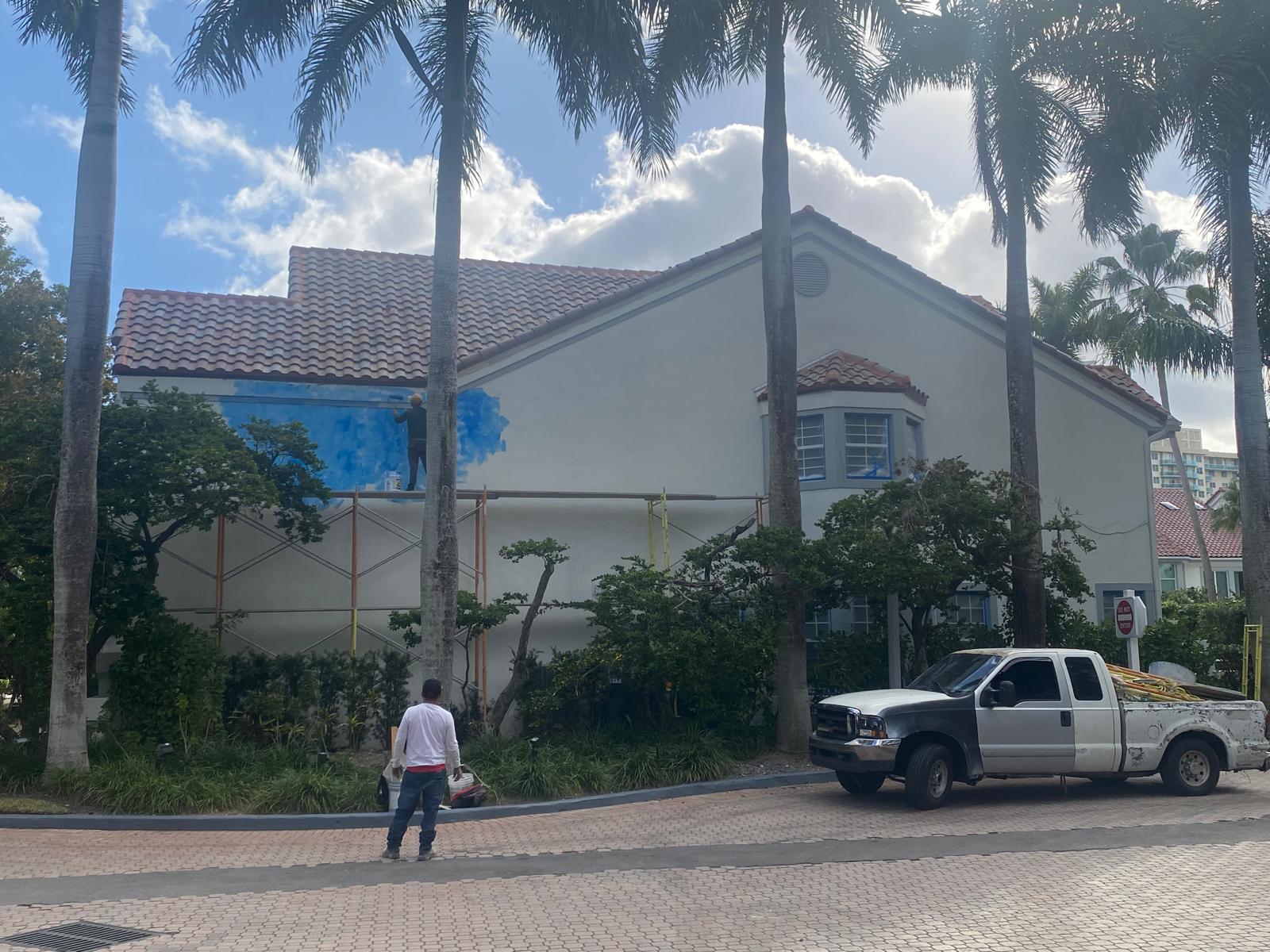 Professional painter applying blue exterior paint on the stucco wall of a Fort Lauderdale home